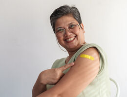 A smiling woman points to the adhesive bandage on her upper arm.